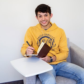Person in a yellow Anoka-Ramsey Community College sweatshirt smiles while holding a pen and writing in a notebook, seated on a gray couch with a small white table.