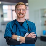 Medical professional in blue scrubs with stethoscope standing confidently in a clinic lobby.