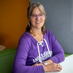 Person sitting at a table, wearing glasses and a purple hoodie smiling at the camera in a brightly colored room.