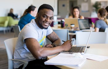 Student smiling with laptop and papers on table.