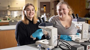 Students in lab setting sitting behind microscopes, looking at camera smiling