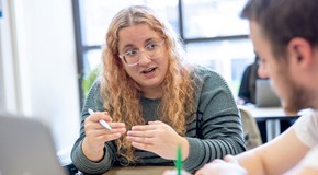 student with glasses and pen in hand talking to another student