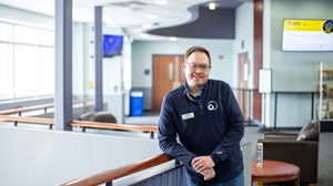 An employee with a badge and logo shirt smiling, standing by a wooden railing with seating areas in the background.